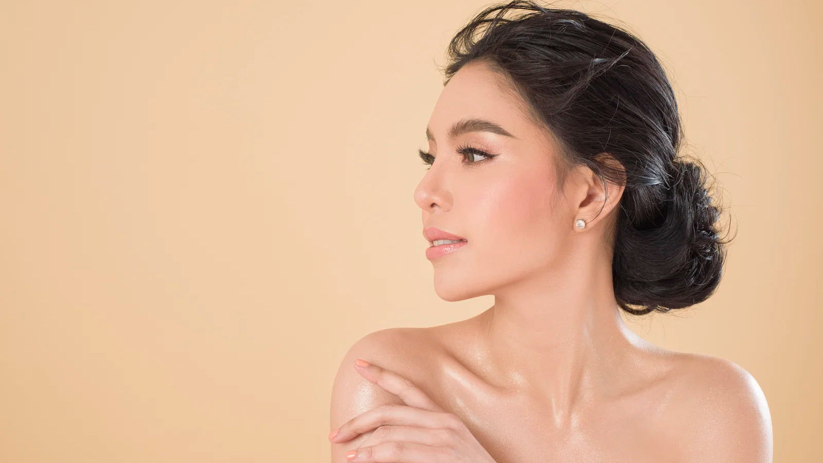 A woman with dark hair styled in an elegant low bun, wearing a small gold stud earring, shown in profile against a solid beige background.
